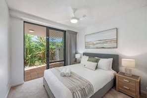A bedroom with a bed, a nightstand, a ceiling fan, a window with a view of greenery, and a painting on the wall. - Pawfect Coastal - Pet friendly, Beachfront, Pool (Marcoola)