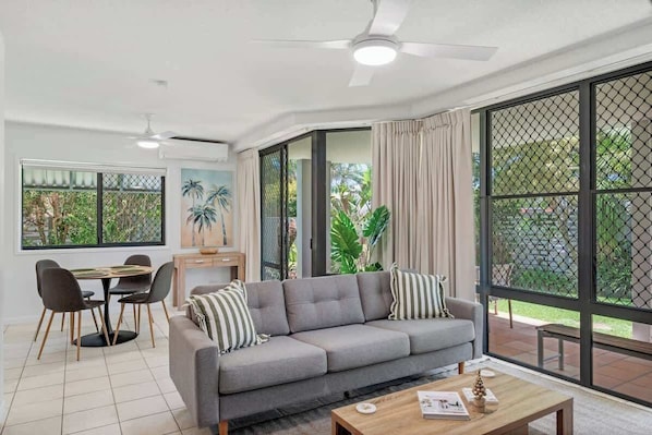 A modern living room with a grey sofa, a dining table with chairs, a ceiling fan, and large windows with curtains. - Pawfect Coastal - Pet friendly, Beachfront, Pool (Marcoola)