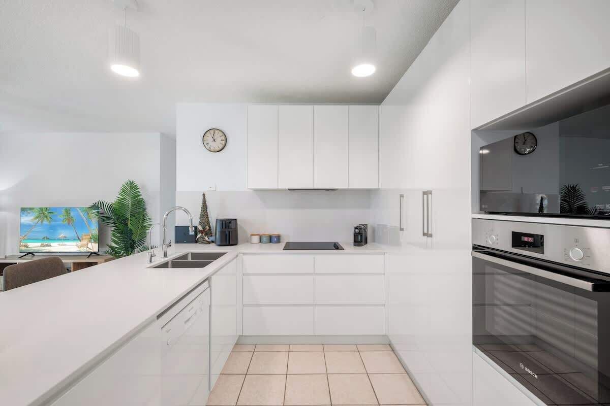 A modern kitchen with white cabinetry, a Bosch oven, a clock, and a television displaying a beach scene.