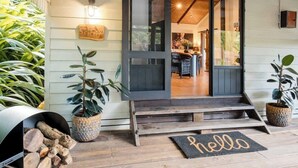A wooden porch with a 'hello' mat, a stack of firewood, and potted plants.