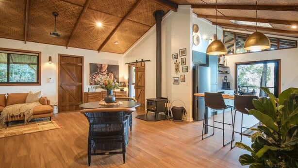A spacious living area with a high ceiling, wooden flooring, a sofa, a dining table, and a kitchen area with bar stools.