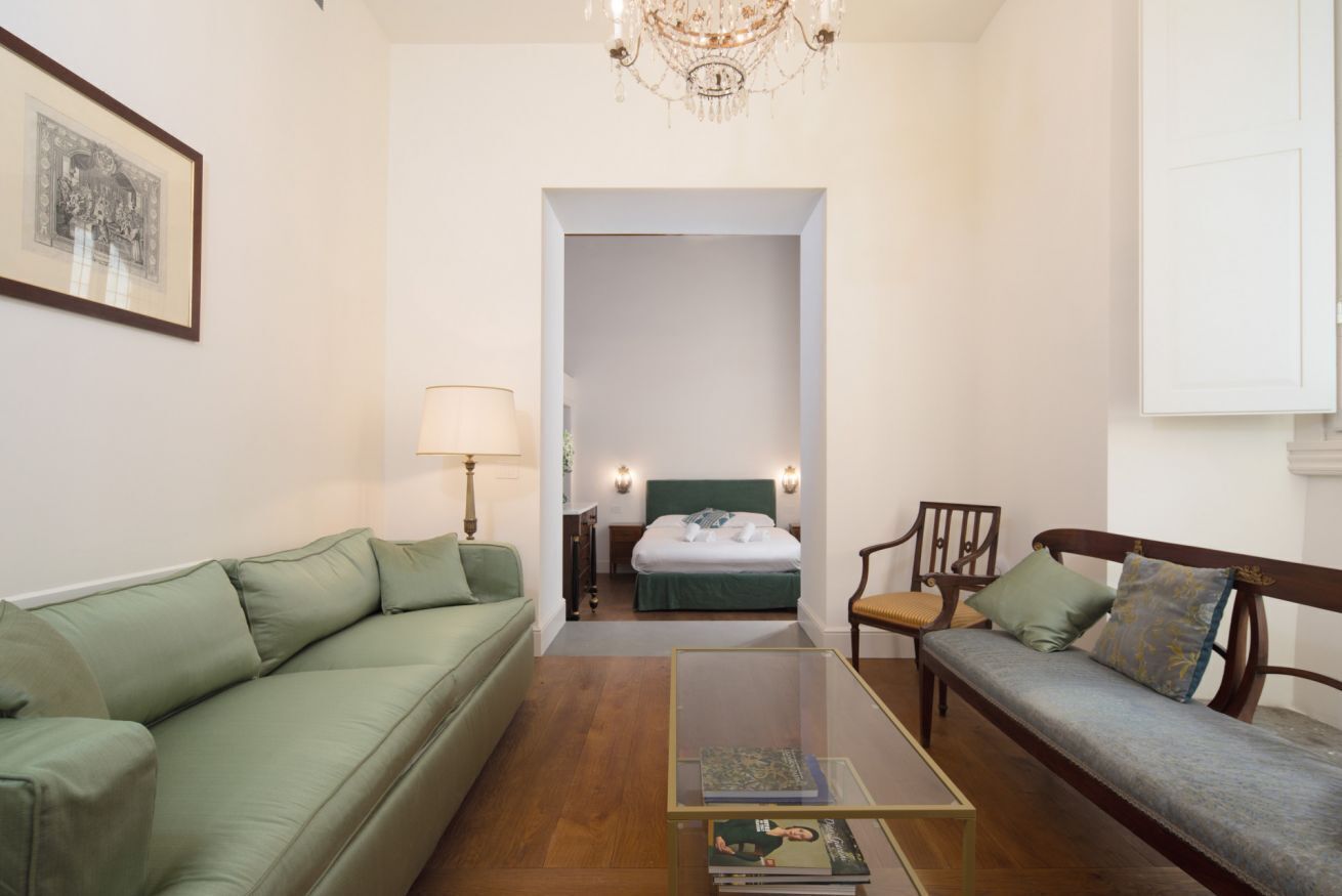 A room with a green sofa, a wooden bench, a glass coffee table, a bed with a green headboard, and a chandelier.