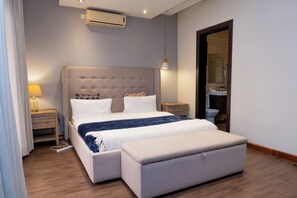A modern bedroom with a tufted headboard, a bedside table with a lamp, a bench at the foot of the bed, and an air conditioning unit. - Fallsway Palms (Lusaka)