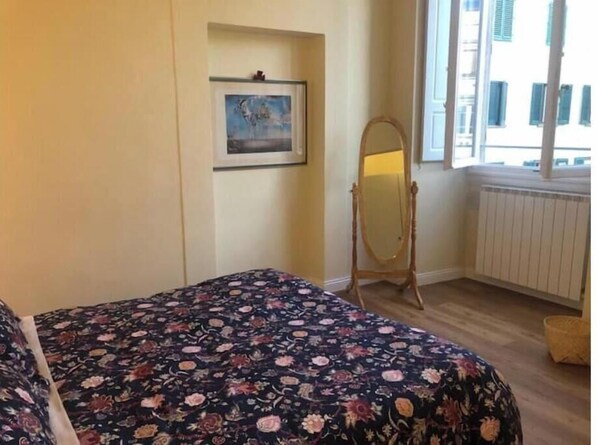 Une chambre à coucher comprenant un lit, une commode, un miroir et une fenêtre donnant sur des immeubles.