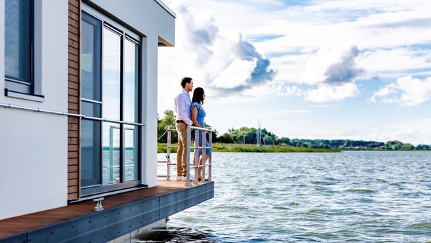 A couple standing on a deck of a modern building with large windows overlooking a body of water. - Kranichnest Schwimmhaus - Kranichnest Schwimmhaus: lake view, terrace, wood-burning stove (Ribnitz-Damgarten)