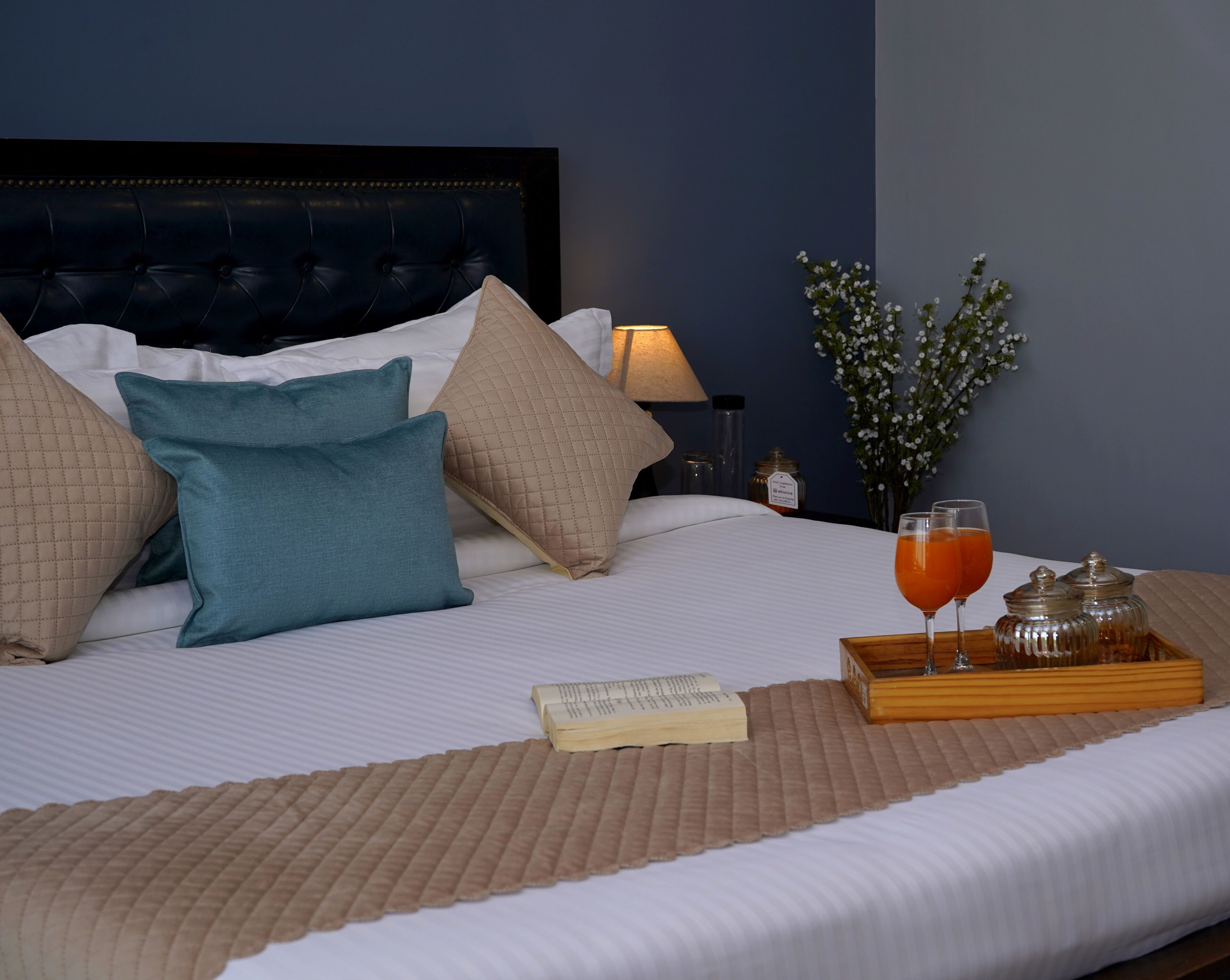 A neatly made bed with pillows, a bedside table with a lamp, a tray with glasses and jars, and a vase with flowers.