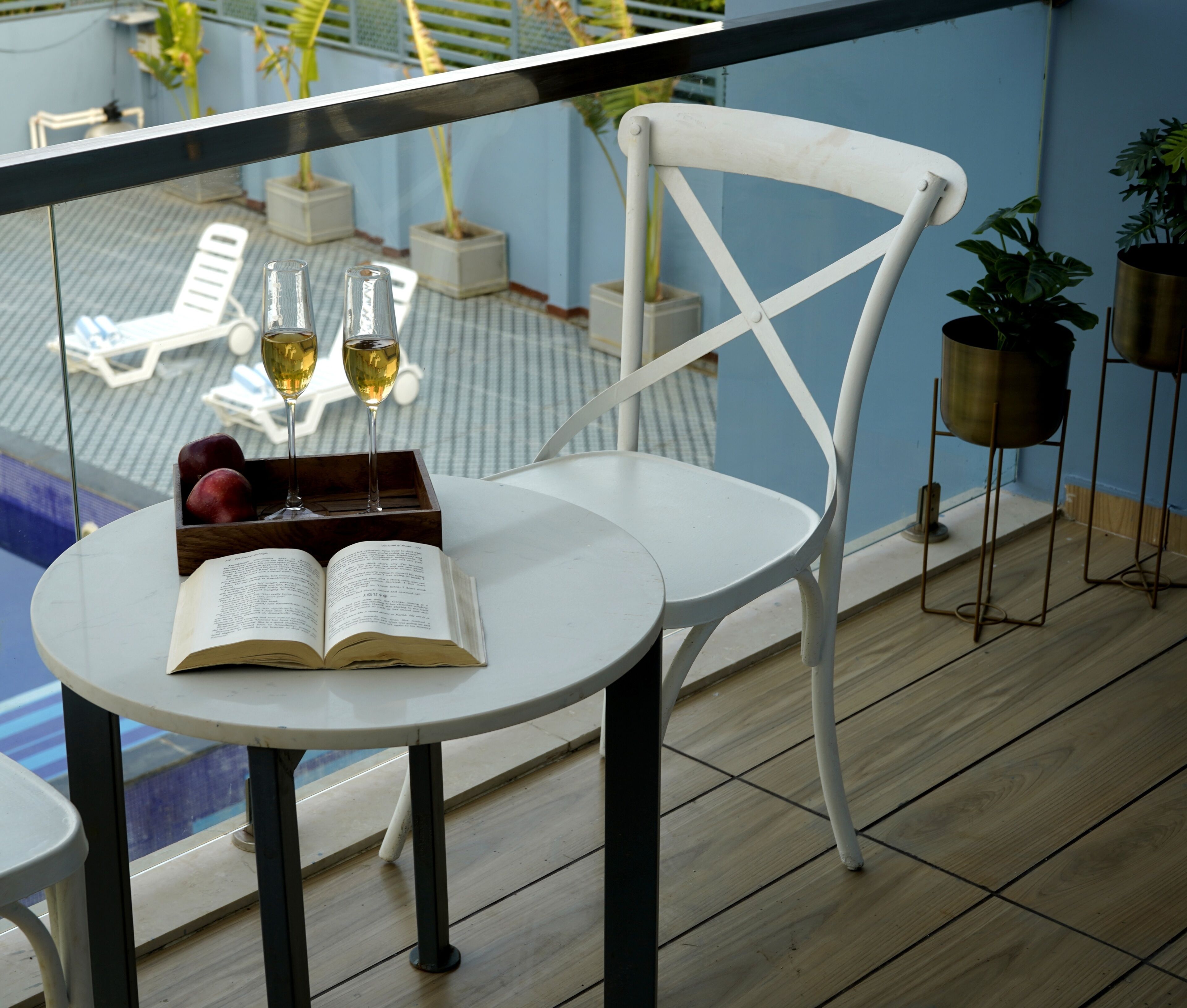 A balcony with a white chair, a small table with an open book and a glass of wine, and potted plants.