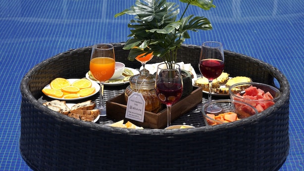 Ontbijt bij het zwembad, met een rieten dienblad, fruit, drankjes en een plant.