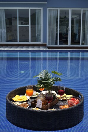 A poolside breakfast setup with a wicker tray, fruits, beverages, and a plant. - ELIVAAS Bluebell Villa 2 BHK Villa (Udaipur)