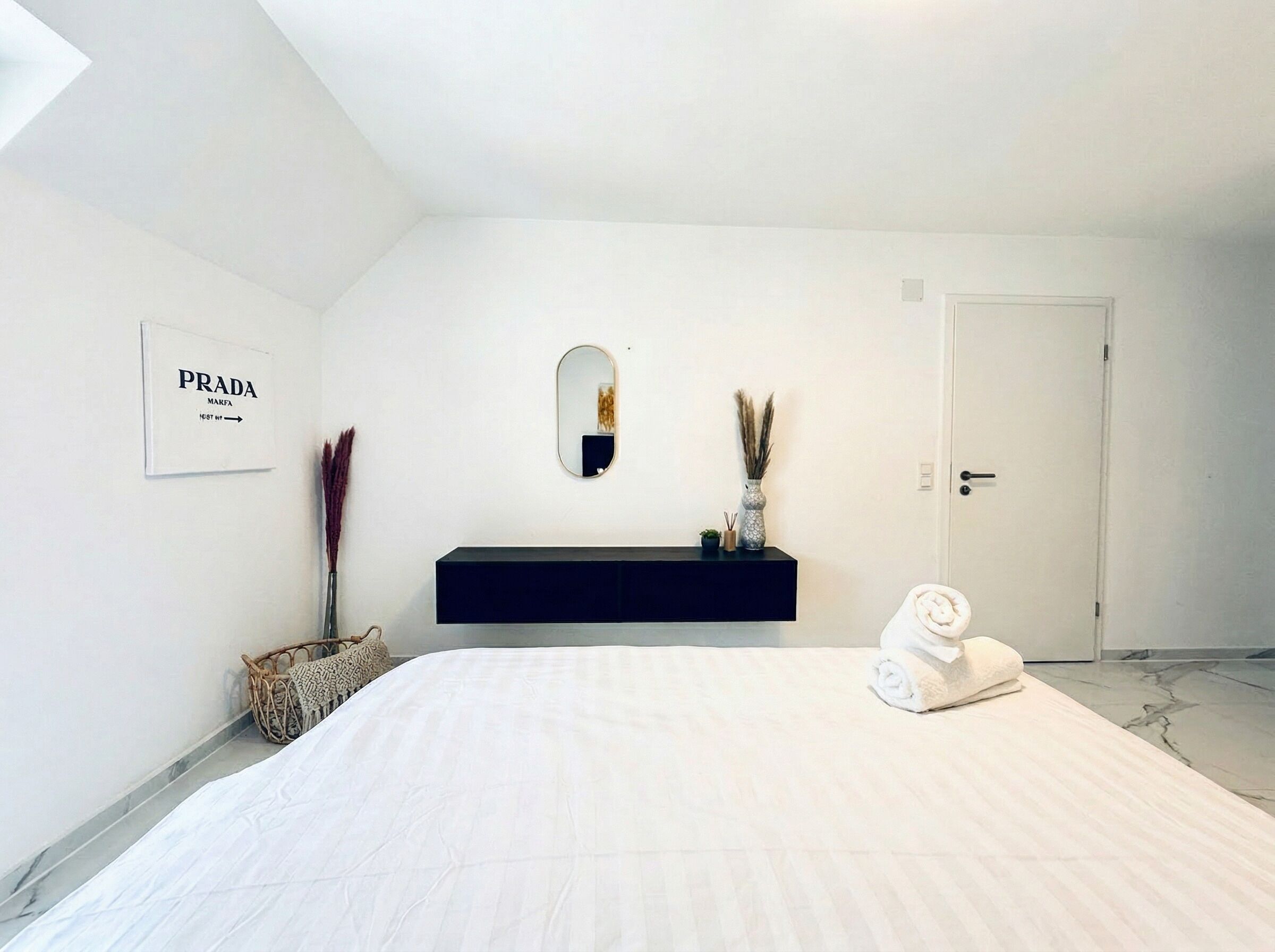A minimalist bedroom with a bed, a dark console table, a mirror, and a door.