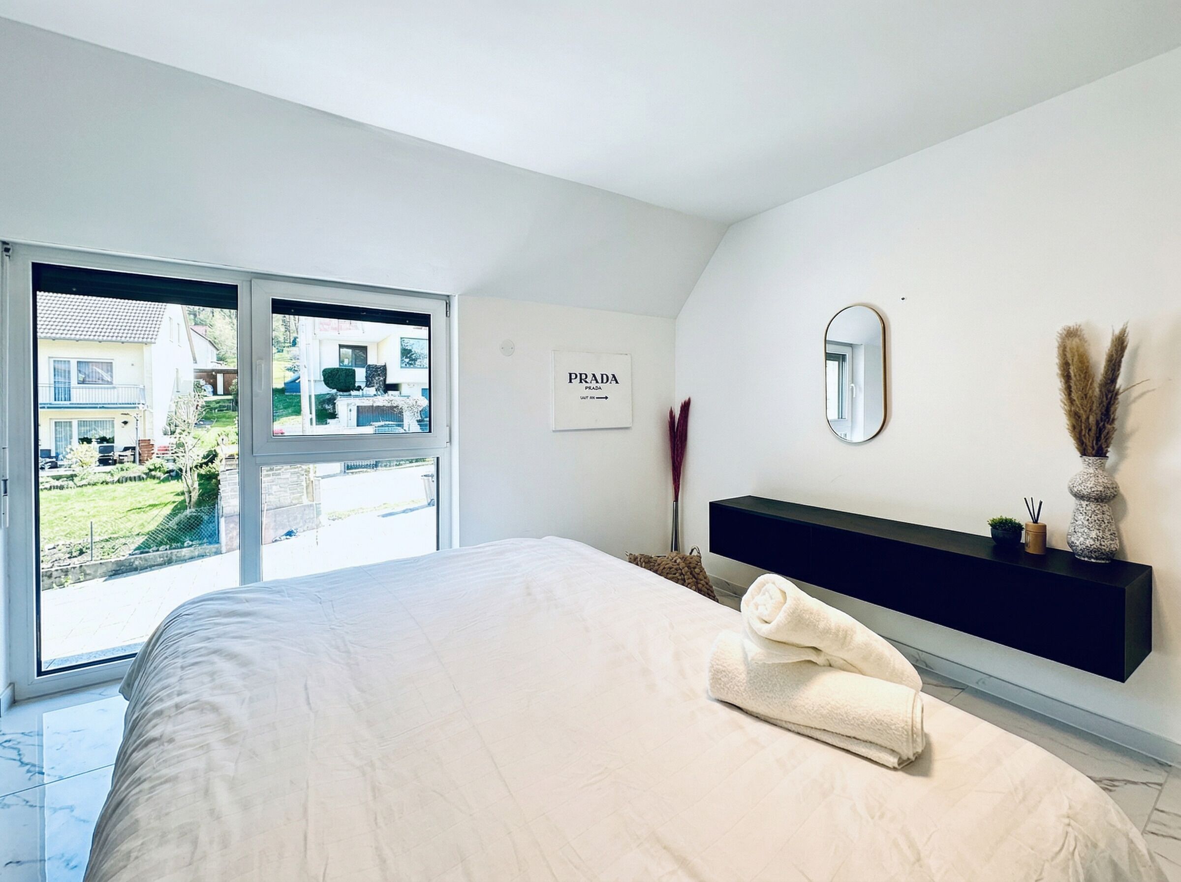 A bedroom with a bed, a bedside table, a mirror, a wall-mounted TV, and a window with a view of a house and garden.