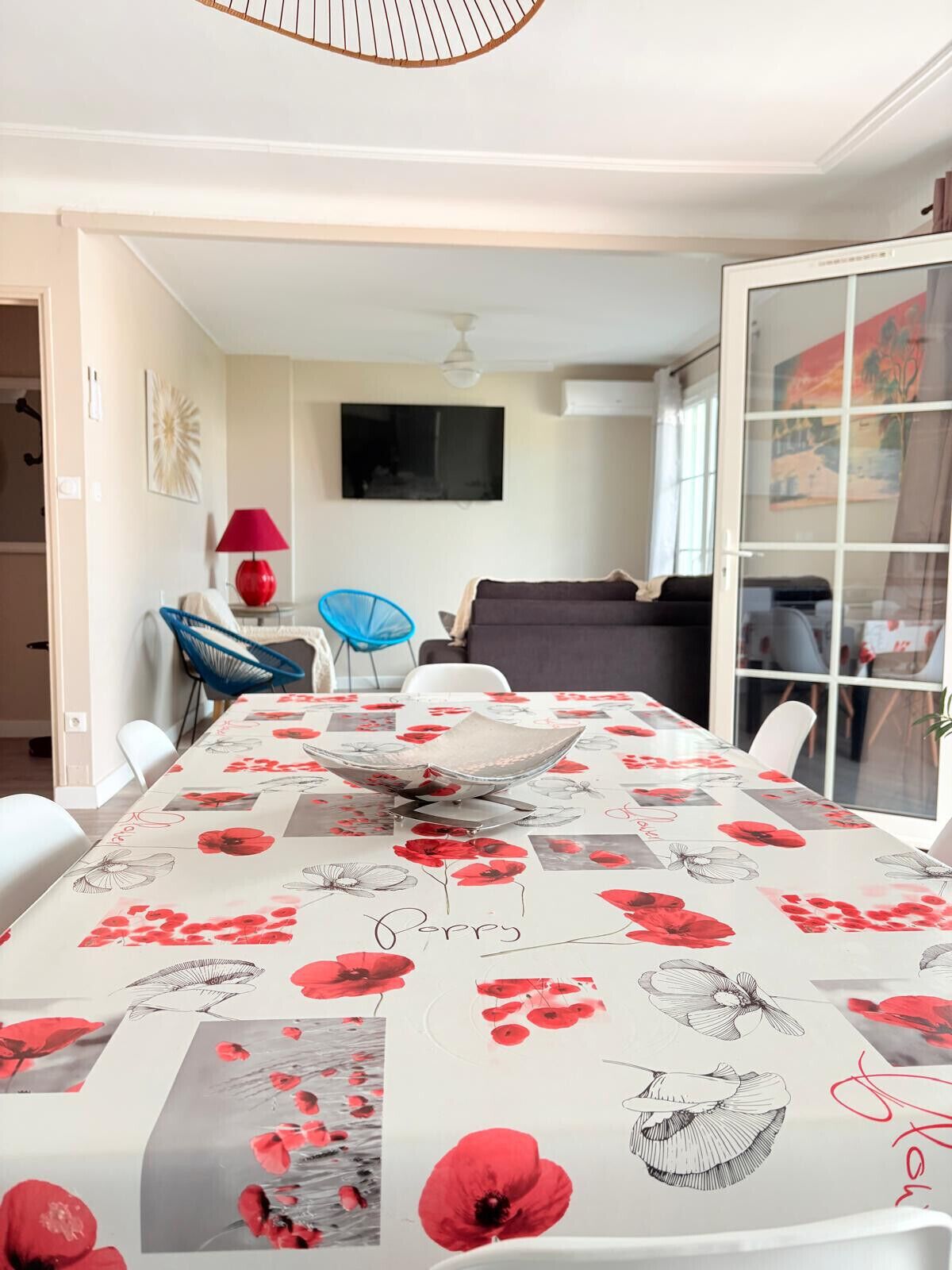 Un coin repas avec une table recouverte d’une nappe à motifs floraux, un téléviseur fixé au mur et une porte-fenêtre coulissante donnant sur un balcon.