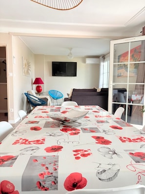 A dining area with a table covered in a floral tablecloth, a television mounted on the wall, and a sliding glass door leading to a balcony. - St Cyprien superb luxury 3-bedroom apartment (Saint-Cyprien)