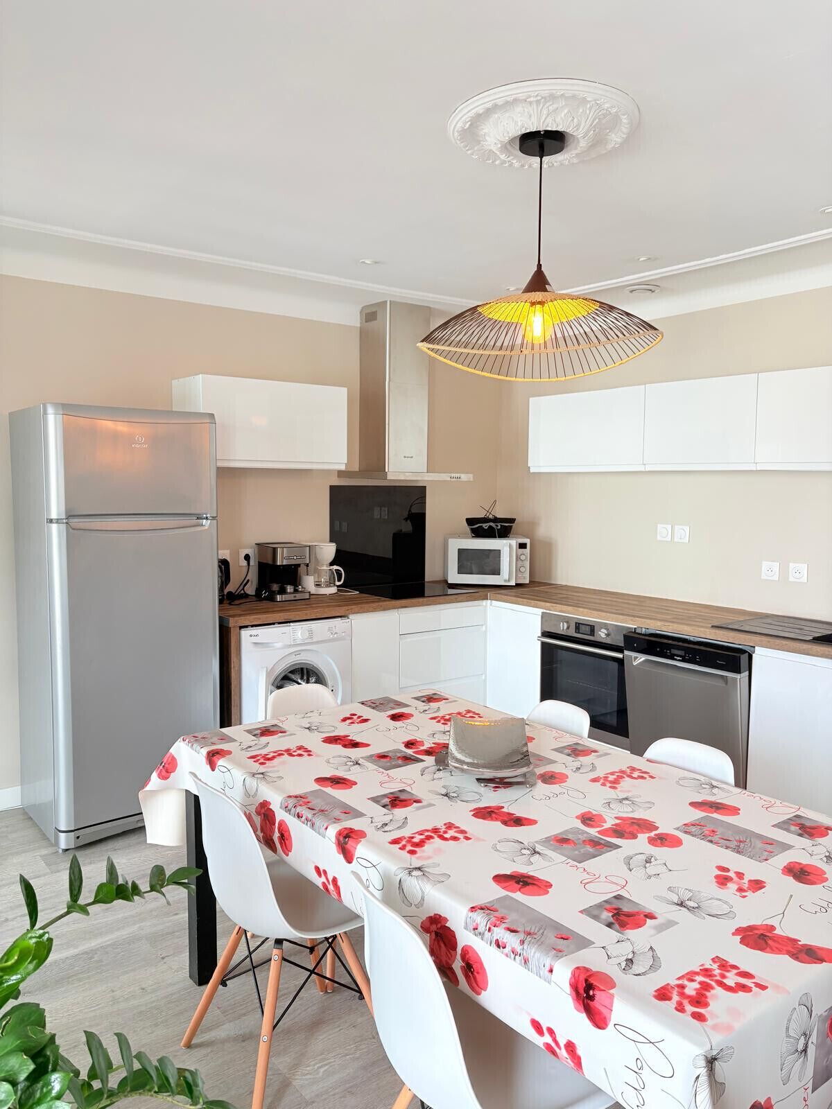 Een compacte keuken met een koelkast, magnetron, oven en vaatwasser. Er is een eettafel met stoelen en een decoratieve plafondlamp.