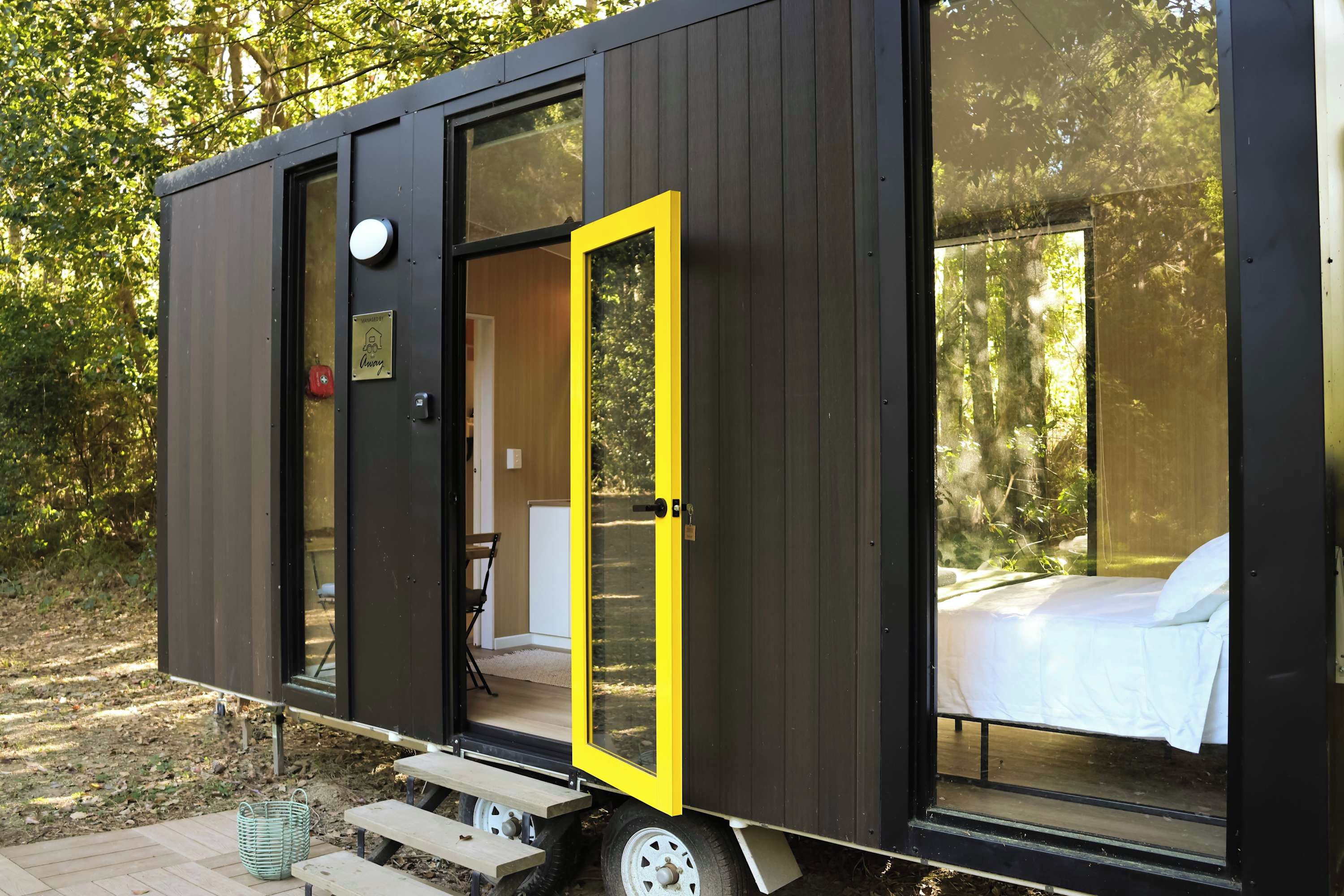 A compact, modern cabin with a sliding door, a small outdoor seating area, and a bed visible through a large window.
