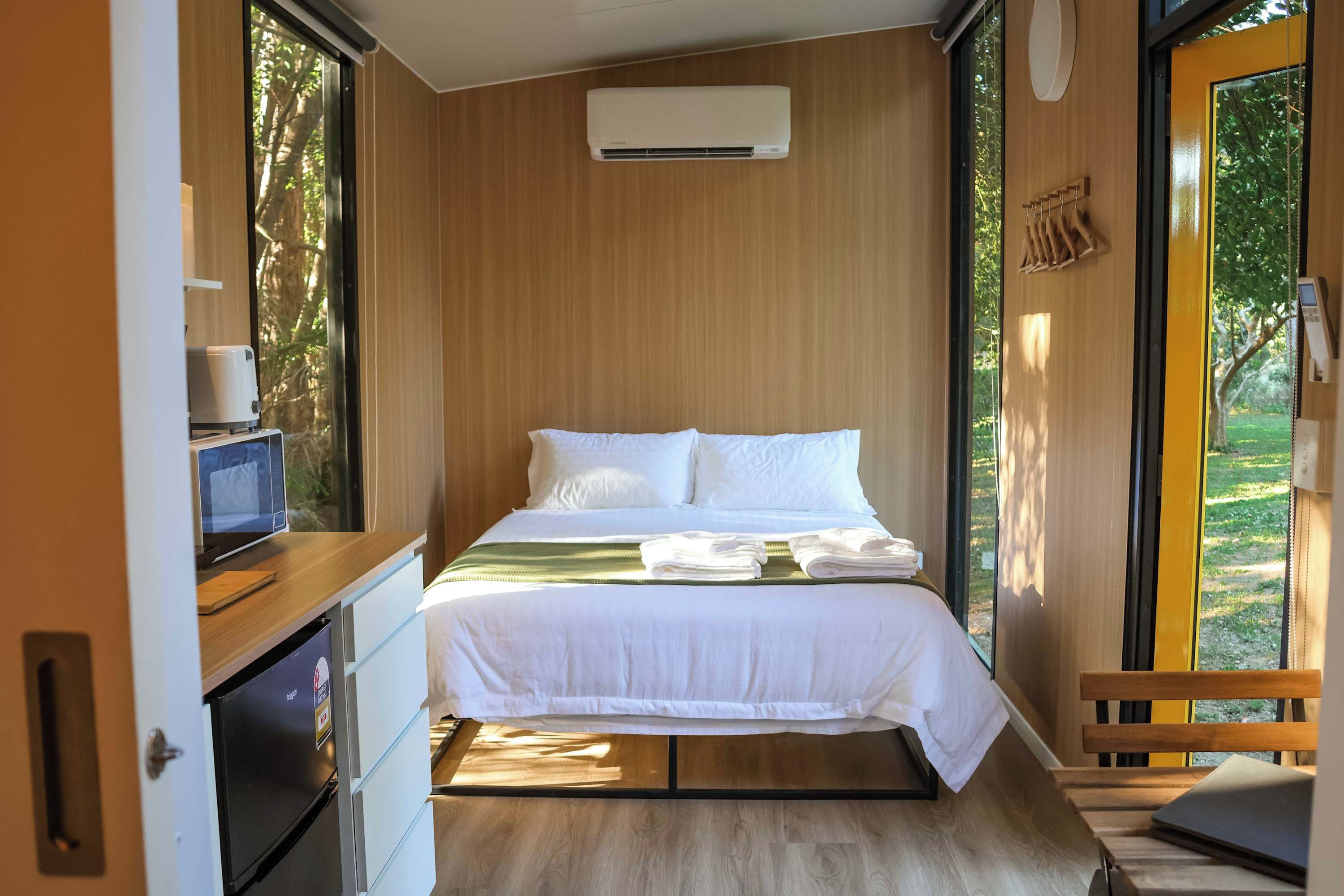 A compact bedroom with a bed, a small desk with a computer, an air conditioning unit, and a view of greenery through the window.