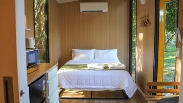 A compact bedroom with a bed, a small desk with a computer, an air conditioning unit, and a view of greenery through the window.