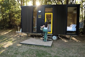 Une petite maison mobile moderne et fonctionnelle, dotée d’une petite terrasse, d’une seule fenêtre visible et d’une porte donnant sur un intérieur lumineux.