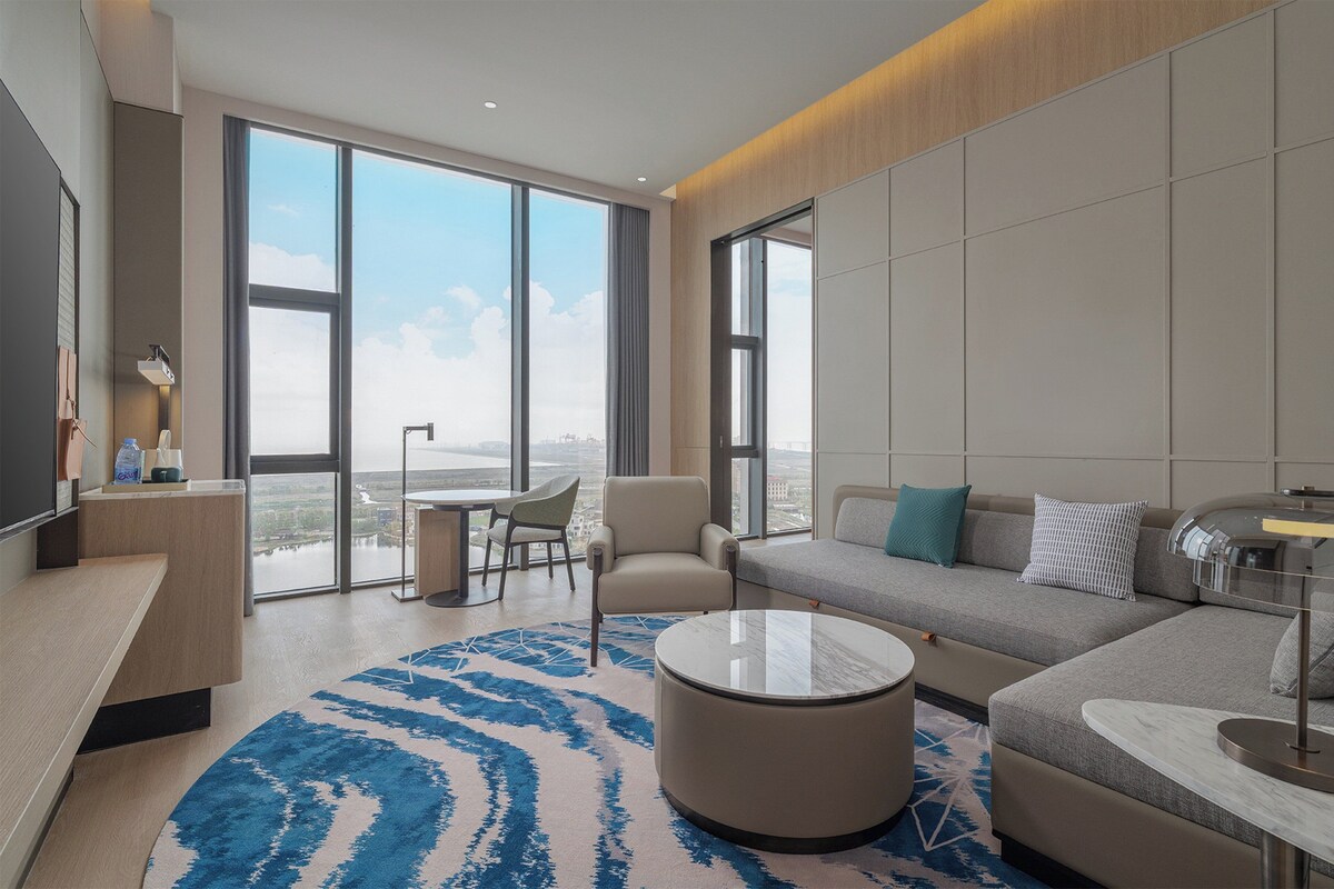 Quarto de hotel moderno com uma janela grande, um sofá, uma mesa de centro redonda e uma escrivaninha.