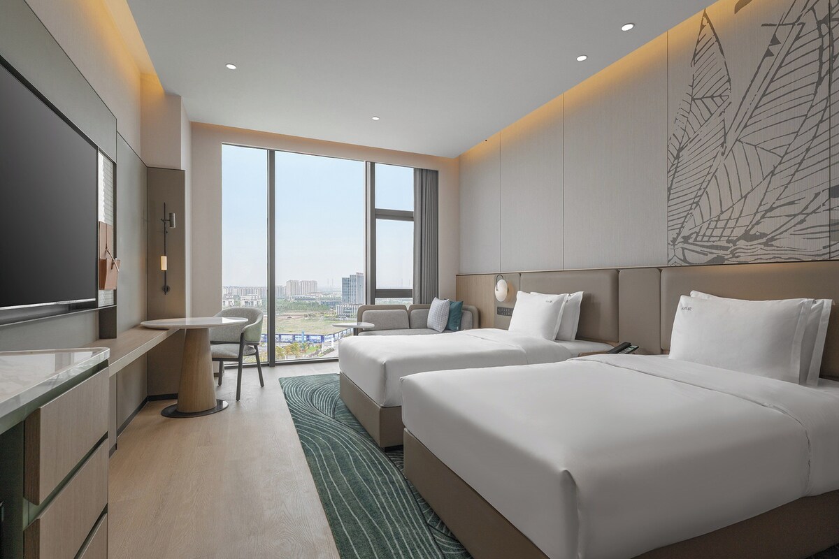 Quarto de hotel com duas camas, uma escrivaninha, uma cadeira e uma janela grande com vista para a cidade.