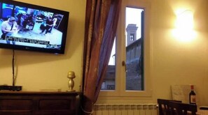 Ein Zimmer mit an der Wand montiertem Fernseher, einem Fenster mit Vorhängen, einem Heizkörper und einer Flasche auf einem Tisch.