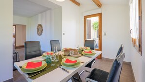 Uma sala de jantar com uma mesa comprida posta para quatro pessoas, cadeiras, um lustre pendurado e vista através de uma porta aberta.