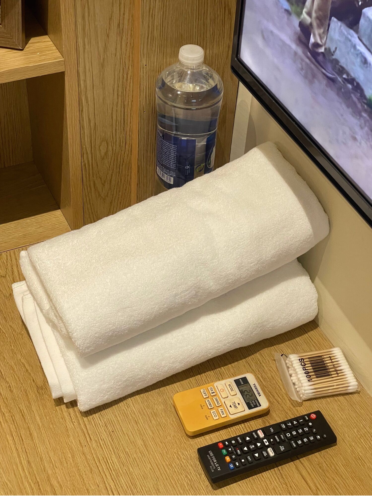 Een netjes gevouwen witte handdoek op een houten oppervlak, een waterfles, een afstandsbediening en een televisiescherm waarop iemand loopt.