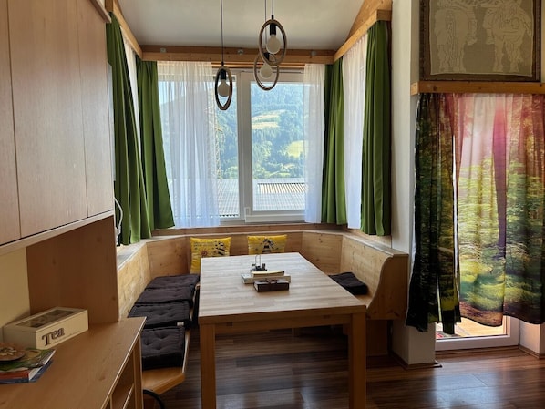 Un cuarto acogedor con una zona de estar con banco de madera, una mesa con libros y una ventana con cortinas verdes.