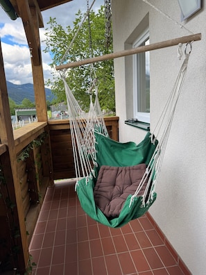 Una silla hamaca verde con un cojín marrón en un balcón con suelo de baldosas y barandillas de madera.