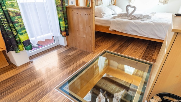 Piso de madeira, um compartimento de vidro com um cavalo, uma cama com roupa de cama branca e uma disposição de almofadas em formato de coração, e uma janela com cortinas.