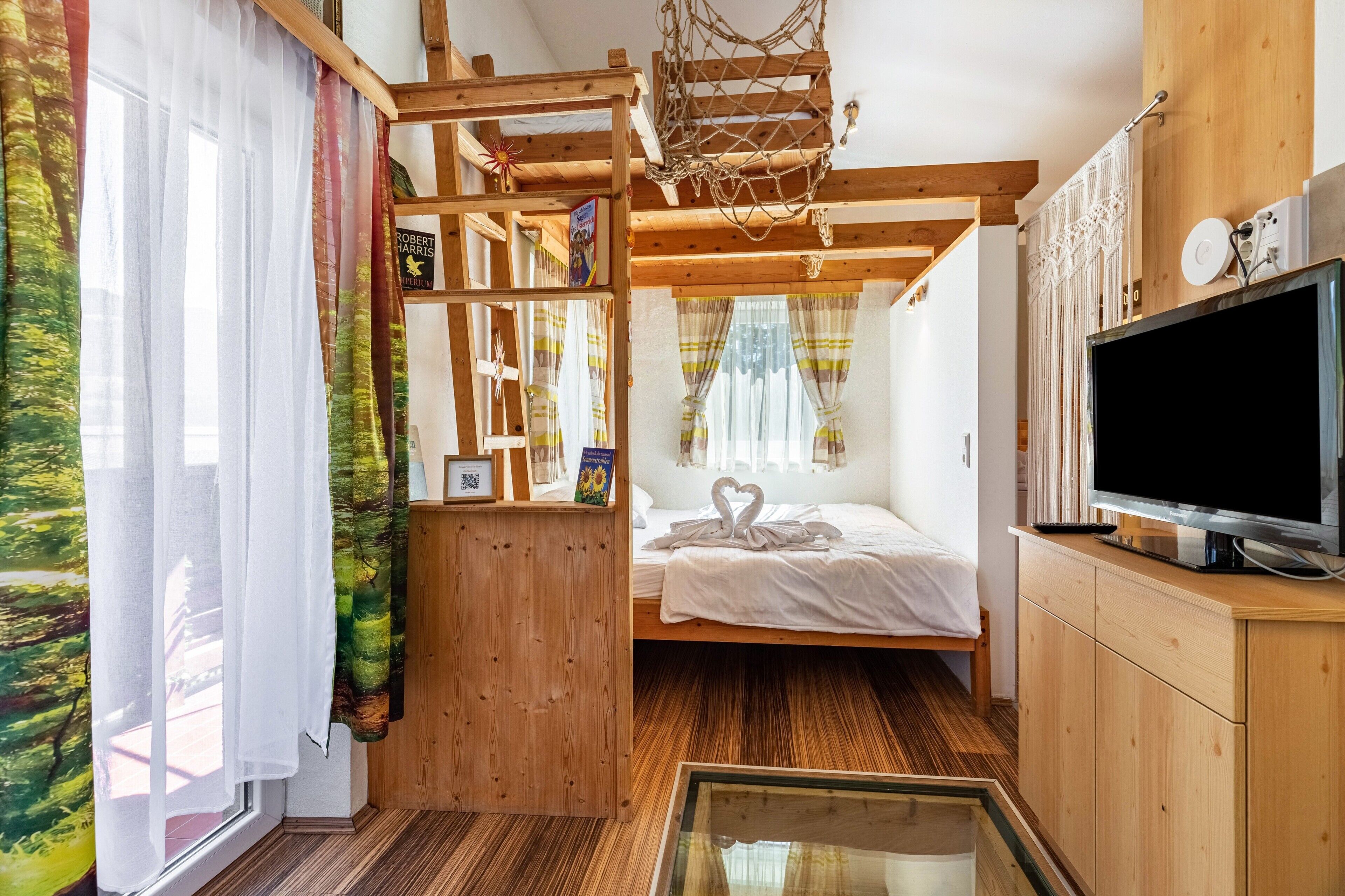 Quarto com cama, televisão, prateleira de madeira, janela com cortinas e estrutura de teto em madeira.