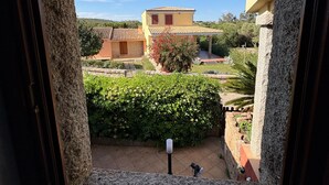 A view through a window with stone frames, a courtyard with a tiled roof building, and a green hedge. - Holiday Home 'Appartamento Baia 12' with Private Garden, Wi-Fi and Air Conditioning (Baia Vignola)