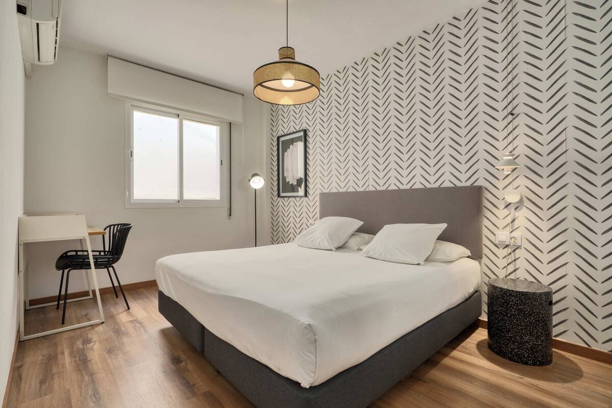 A bedroom with a bed, a desk, a chair, a window, and a wall with a herringbone pattern.