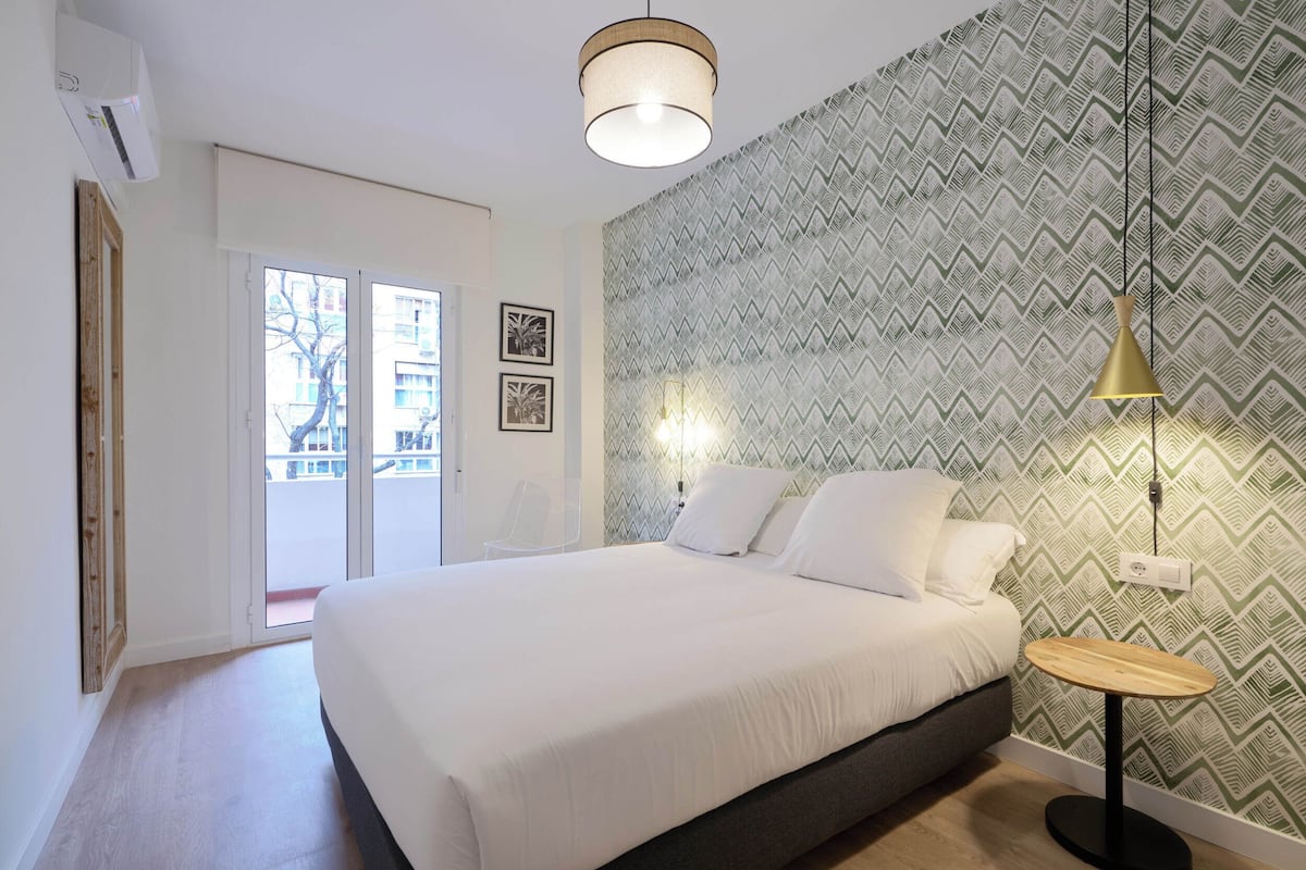 A bedroom with a bed, a small table, a wall-mounted lamp, and a patterned wallpaper.