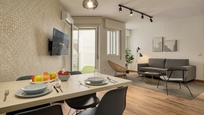 Moderni olohuone, jossa on harmaa sohva, ruokapöytä tuoleineen, seinälle asennettu televisio ja suuri ikkuna.