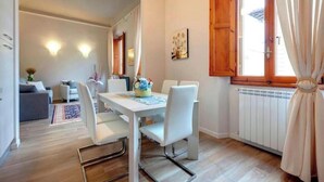 Zona pranzo con tavolo e sedie bianche, una finestra con persiane in legno e un termosifone.