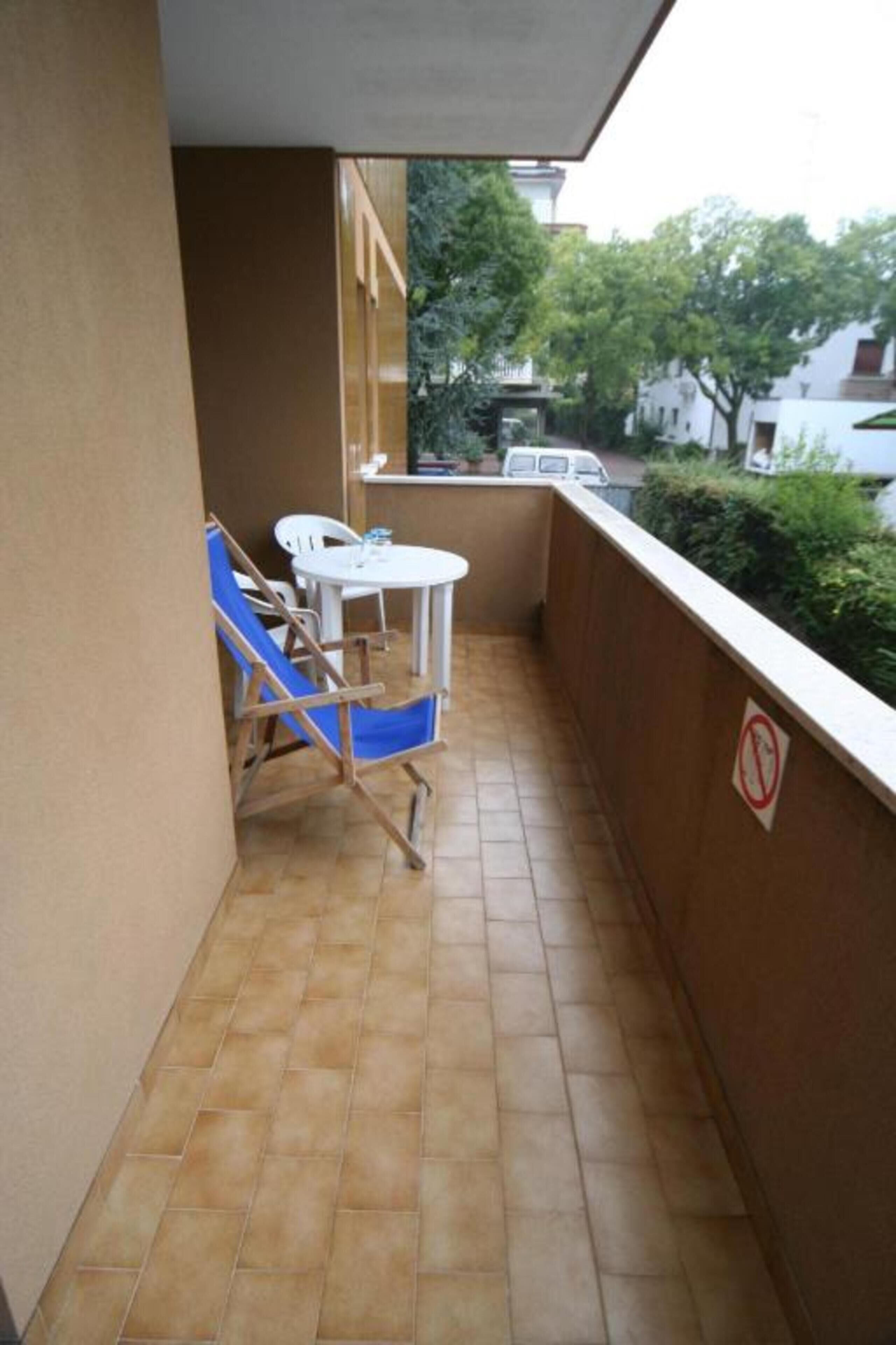 A small balcony with a tiled floor, a white table, two blue chairs, and a white chair.