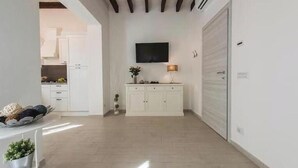 A spacious room with a flat-screen TV mounted on the wall, a white cabinet with drawers, a small table with a lamp, and a large white door. - Little Box House - Modern design and tranquility in the heart of the city (Firenze)