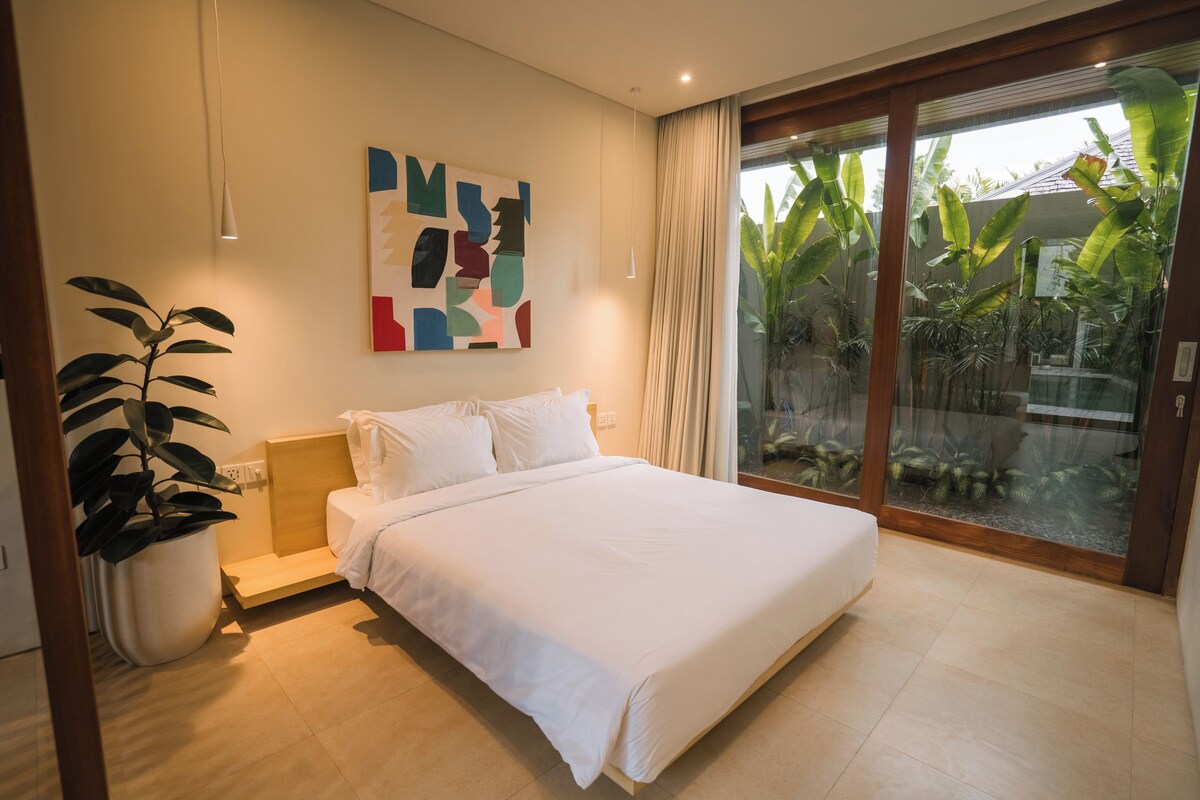 Quarto moderno com cama, planta em vaso, obra de arte na parede e porta de vidro deslizante que dá para uma varanda com plantas.