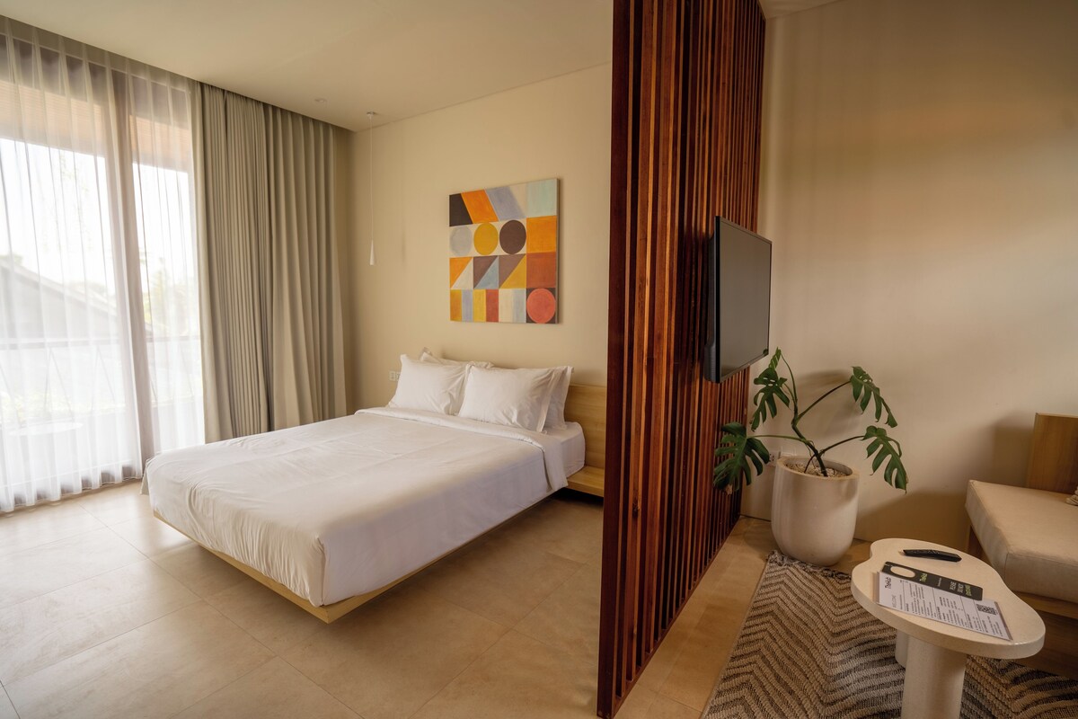 Quarto de hotel com cama, televisão, planta e pequena mesa com cardápio.