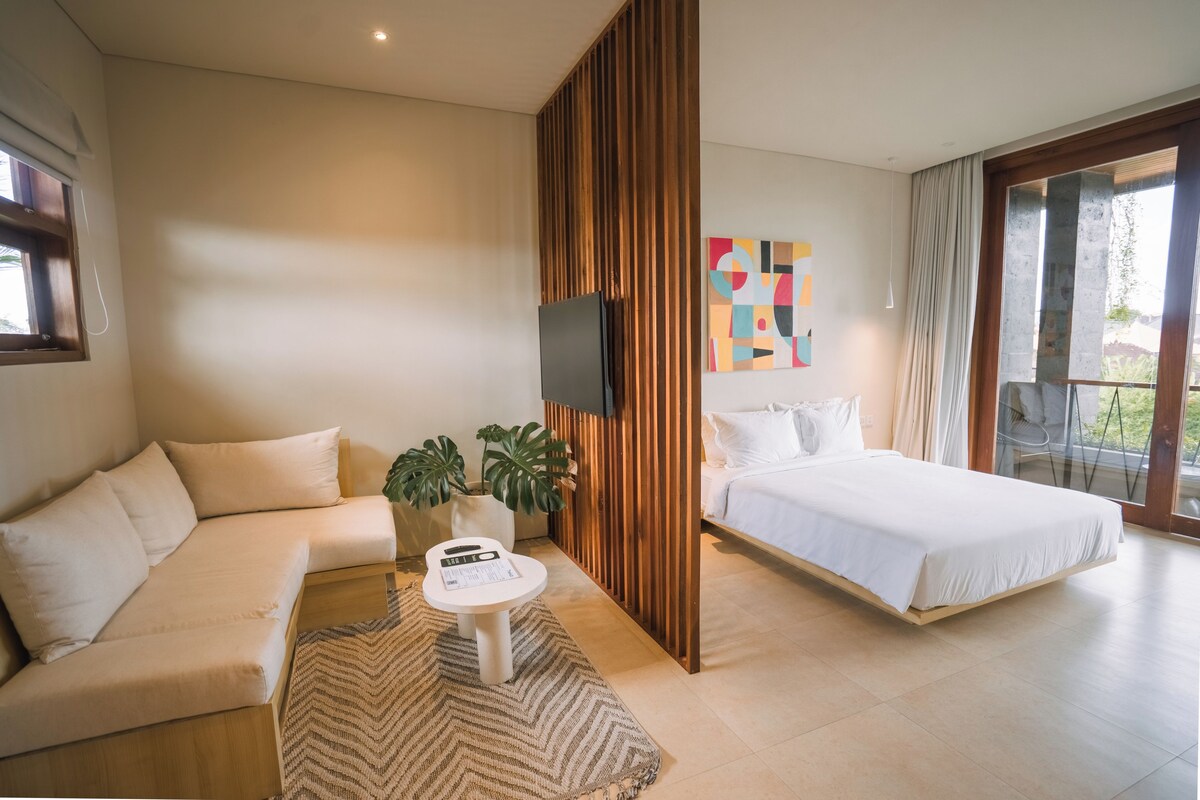 Quarto de hotel moderno com sofá, cama, mesa de centro, televisão e uma janela grande.