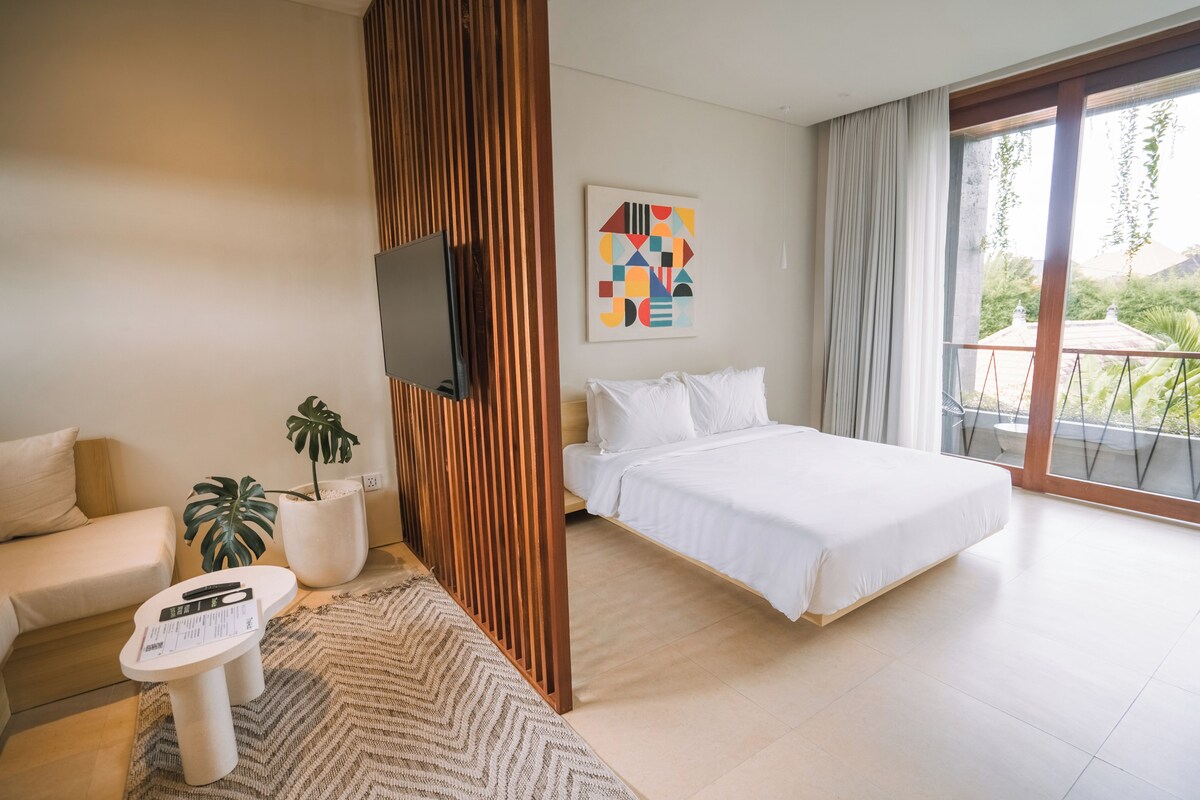 Quarto de hotel com cama, sofá, televisão, planta e pintura.
