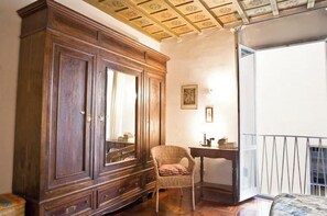 Ein Zimmer mit großem Holzschrank, Korbsessel, kleinem Tisch und einem Balkon mit Geländer.