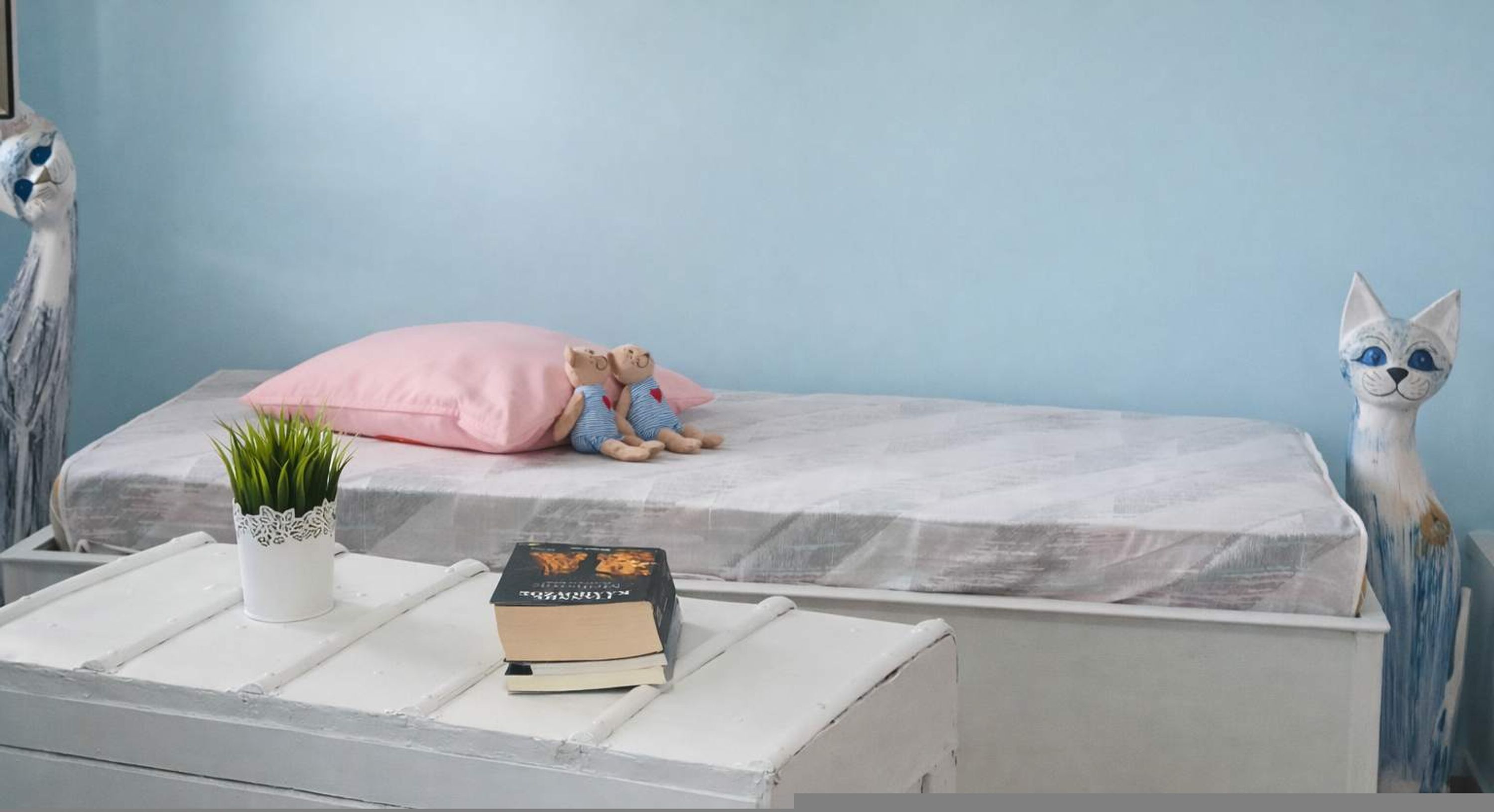 Une chambre à coucher avec un lit, une petite table de chevet ornée d’une plante et de quelques livres, et une statue de hibou décorative.