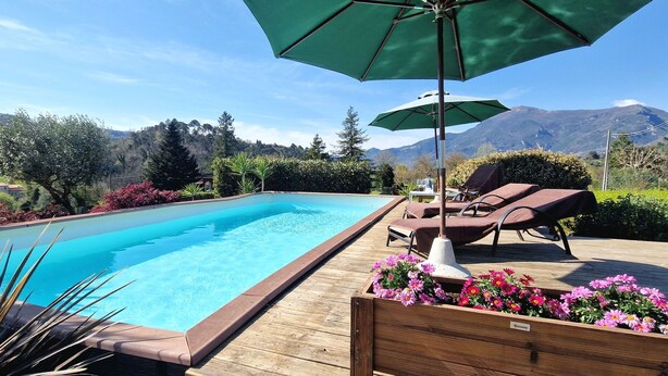 Uma piscina com espreguiçadeiras e um guarda-sol, cercada por um deck de madeira e canteiros de flores, com montanhas ao fundo.