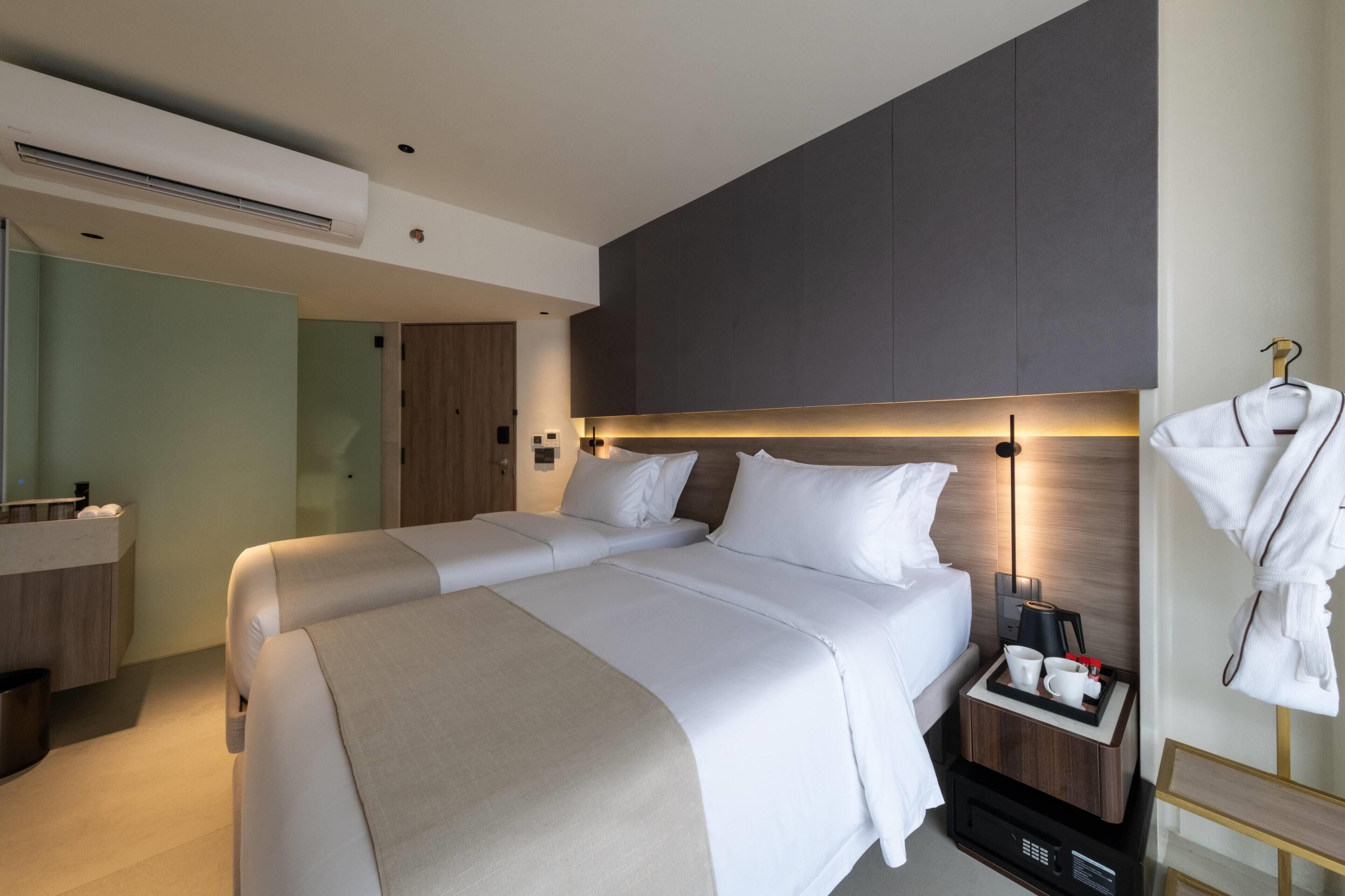 Une chambre d’hôtel moderne avec un grand lit, une table de chevet sur laquelle se trouve un plateau avec des tasses, un peignoir suspendu à un crochet et un grand miroir.