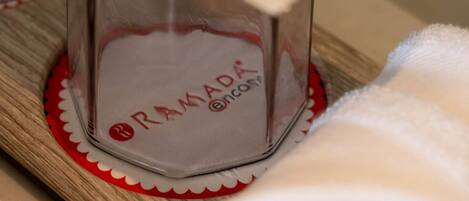 A glass container with the text 'RAMADA' and 'oncore' on it, placed on a wooden surface with a red and white patterned coaster.