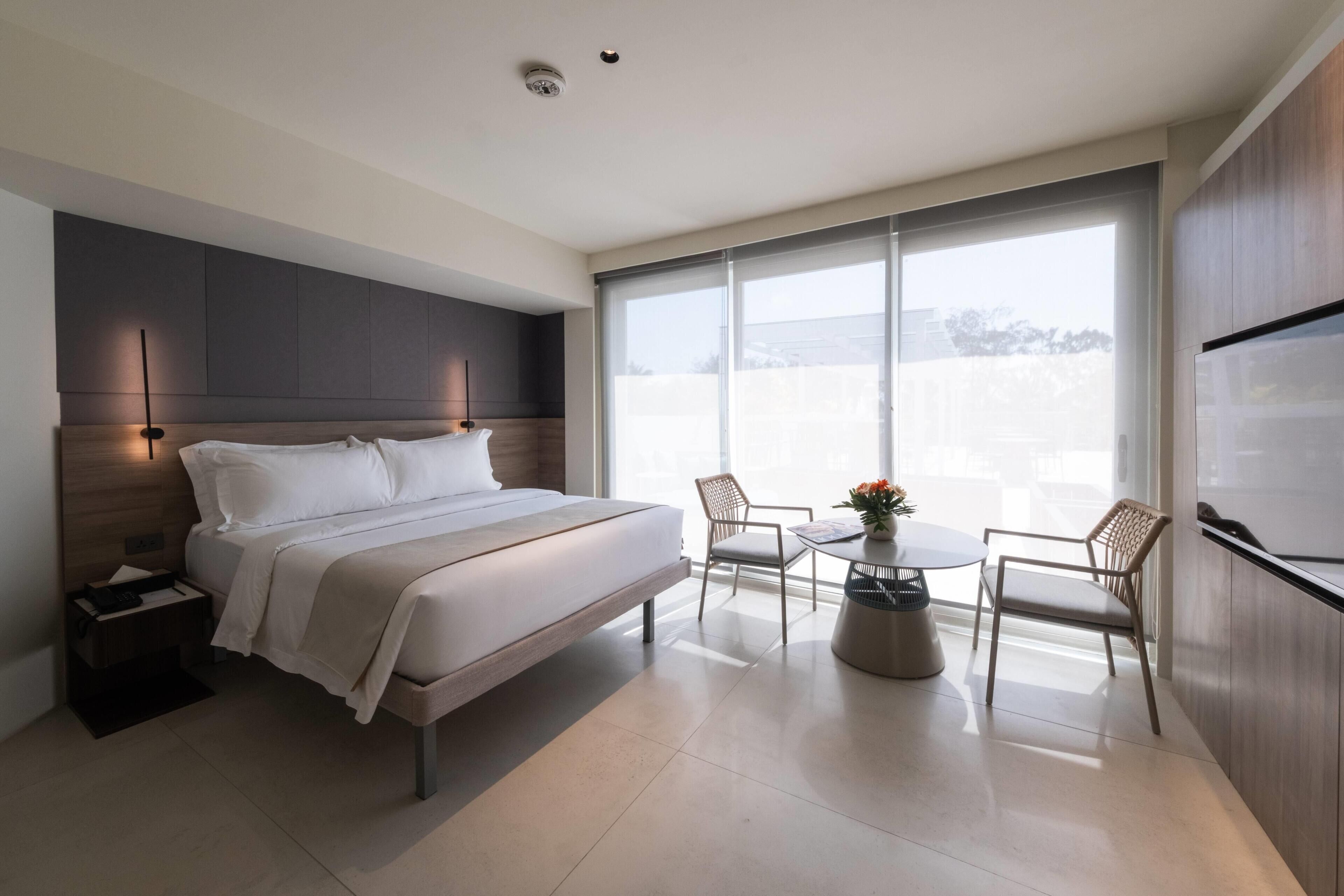 A modern hotel room with a large bed, a bedside table, a small table with chairs, and a large window offering a view.
