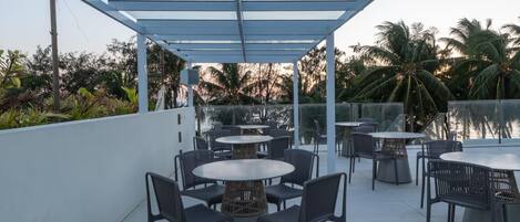 Un patio al aire libre con mesas y sillas redondas, barandilla de vidrio y vistas a palmeras.