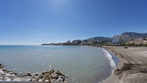 Paesaggio costiero con spiaggia, costa rocciosa e edifici sullo sfondo.